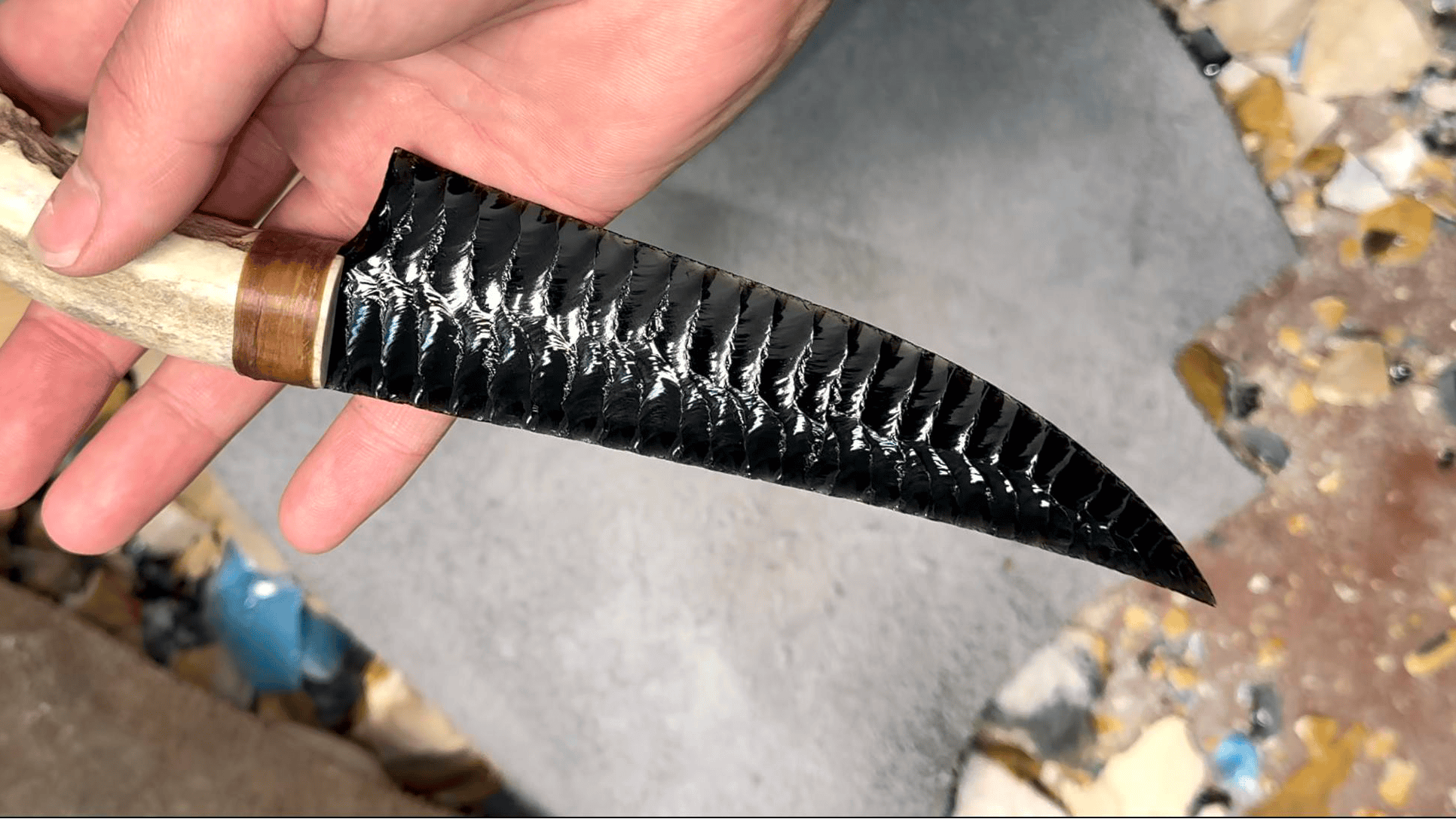 Stoneblade Forge black obsidian curved blade with natural antler handle held in hand, showing hand-knapped texture and glossy finish