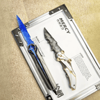 Stoneblade Forge Dagger Stone Blade “Delta Force” — blue crystal-like dagger and grey knapped-style dagger displayed on a “MERCY” spec board in a metal frame.