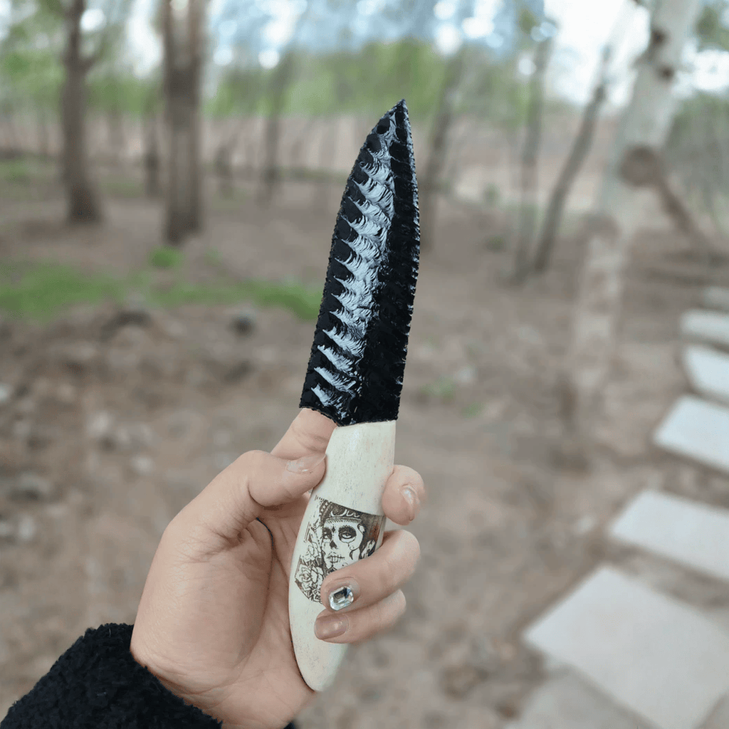 Handheld outdoor shot of the black knapped-style dagger with bone-colored skull-illustrated handle, photographed on a woodland path (Grimwood).