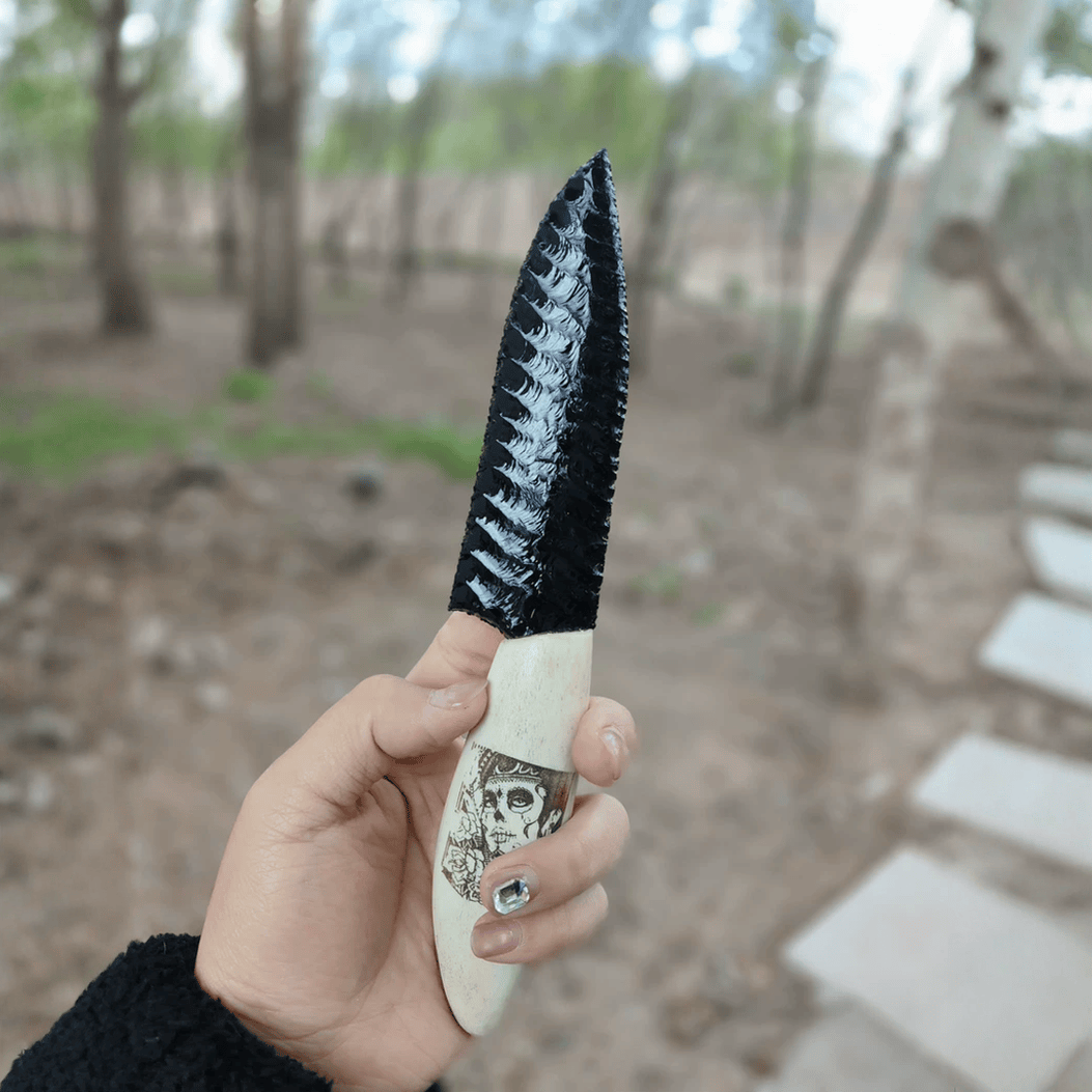 Handheld outdoor shot of the black knapped-style dagger with bone-colored skull-illustrated handle, photographed on a woodland path (Grimwood).