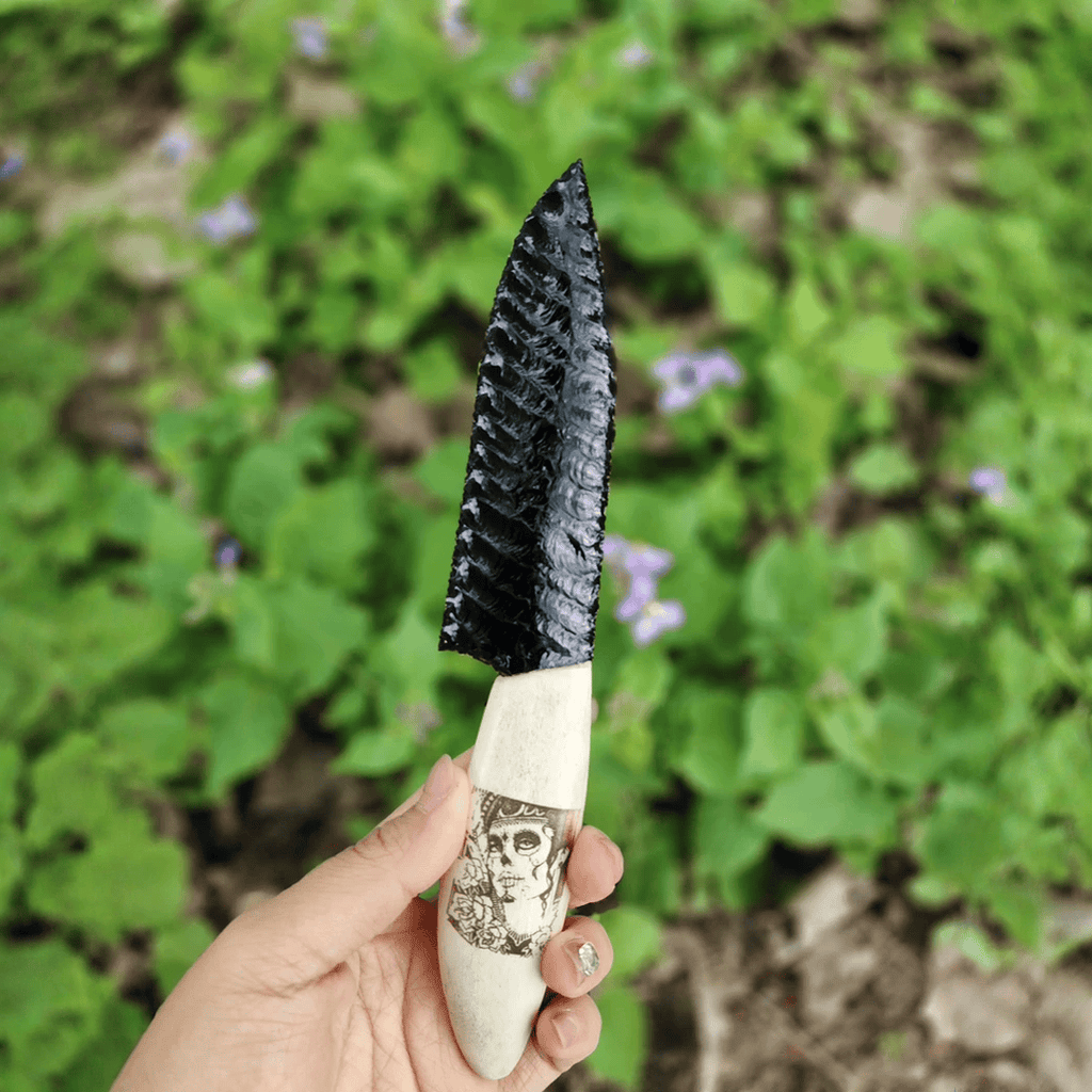 Handheld close-up of the black knapped-style dagger against green foliage, showing the flaked blade pattern and skull-illustrated handle (Grimwood).