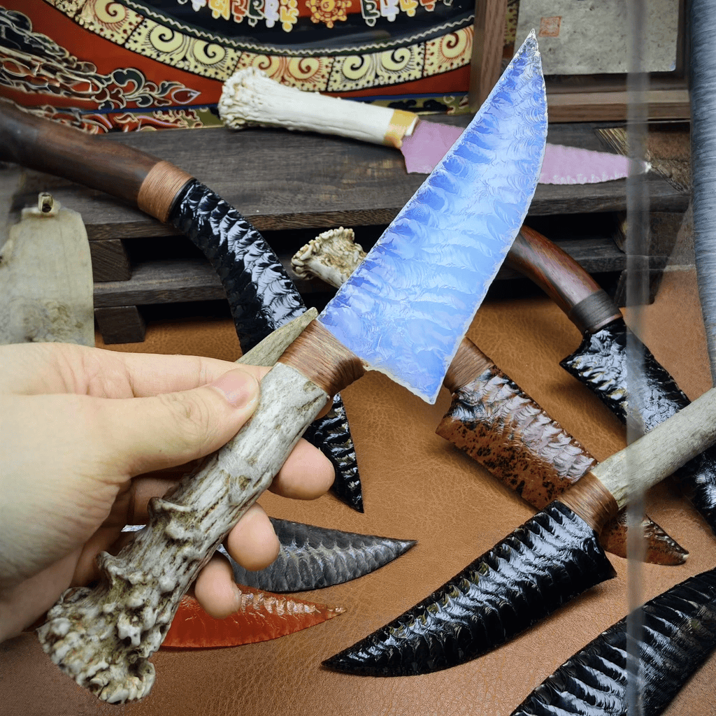 Stoneblade Forge Dagger Stone Blade “Moonwisp” — handheld close-up showing the icy blue blade glow and antler handle, photographed over a display of other stone blades.