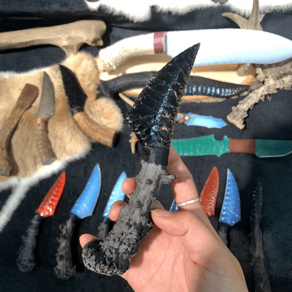 Handheld display of the black knapped-style dagger with rugged dark handle, photographed above a tabletop filled with stone blades and tools (Nightfang).
