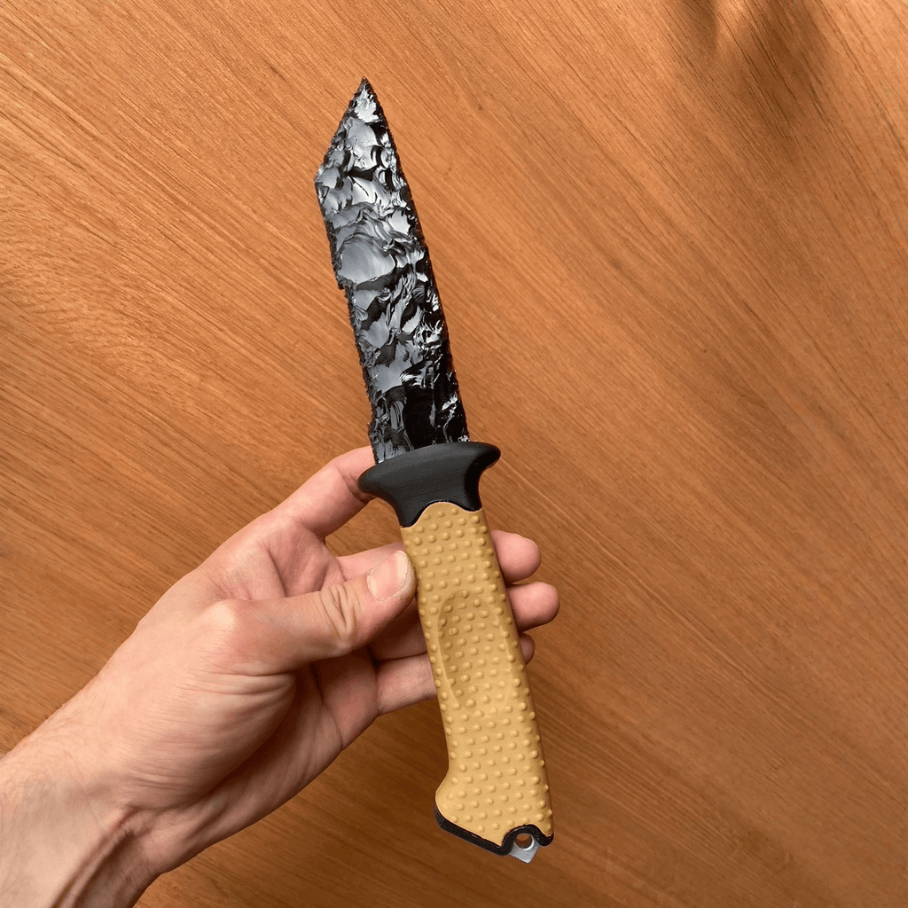 Handheld shot of the Survival dagger, highlighting the knapped black blade texture and tan non-slip handle against a wood background.