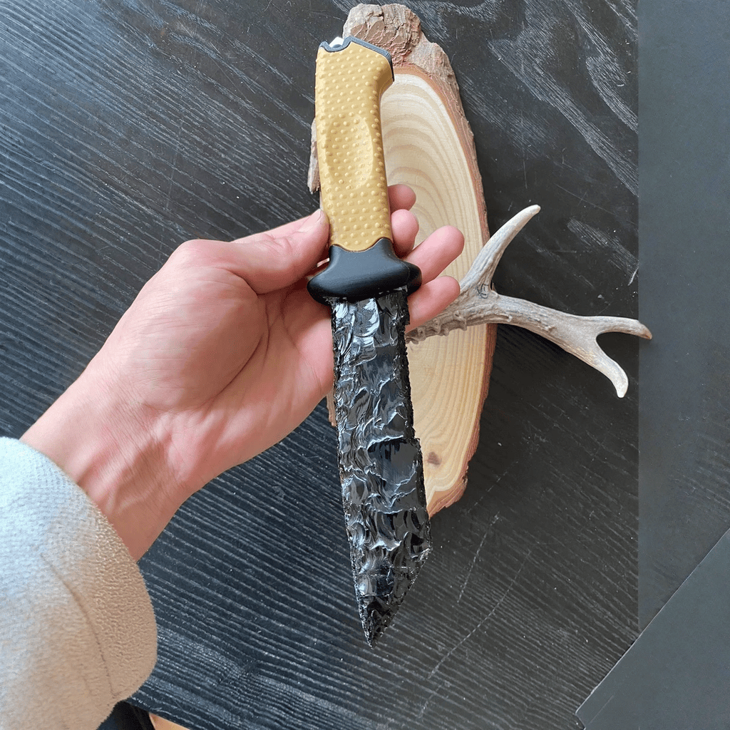 Handheld close-up of the Survival dagger on a dark tabletop with a wood slab and antler prop, showing the blade’s reflective scar pattern and grip detail.