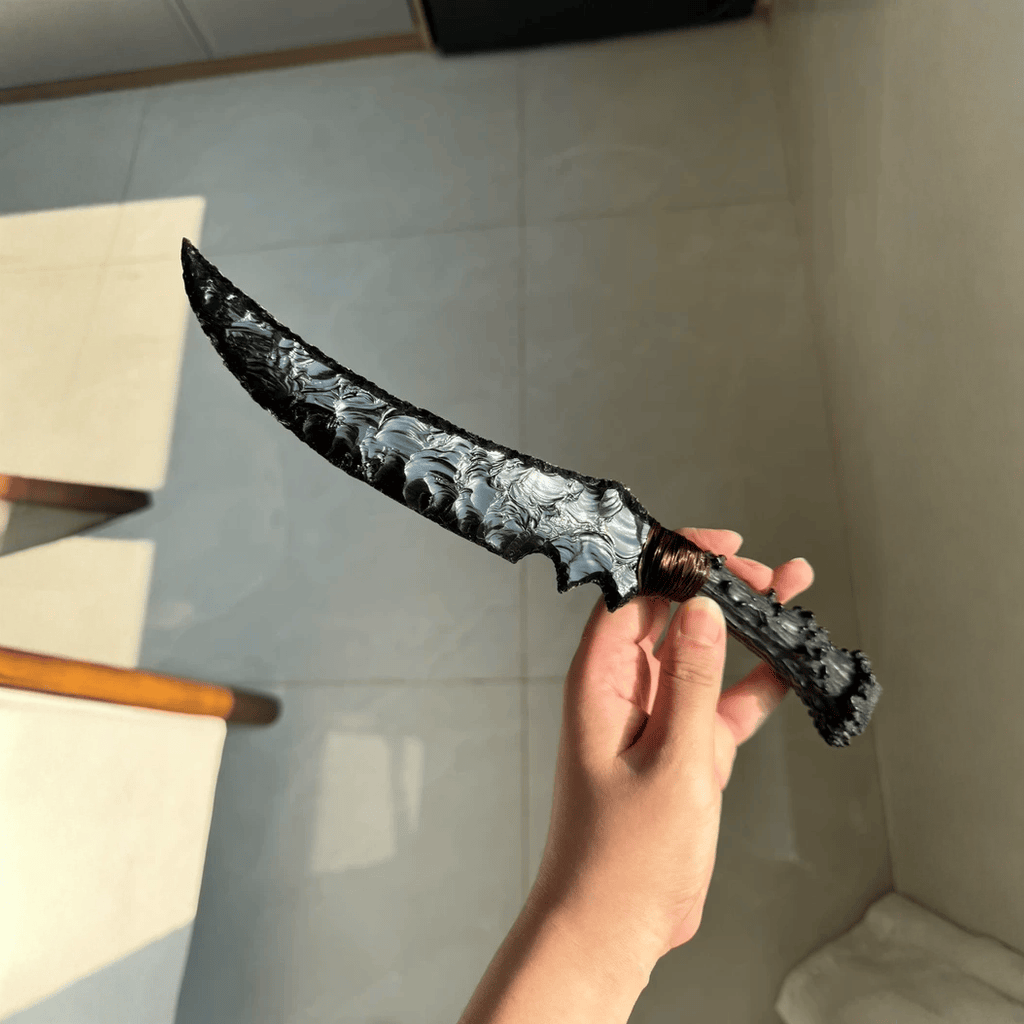 Handheld photo of the curved black knapped dagger showing the reflective flake-scar pattern and copper wire wrap on the handle (Wraith).